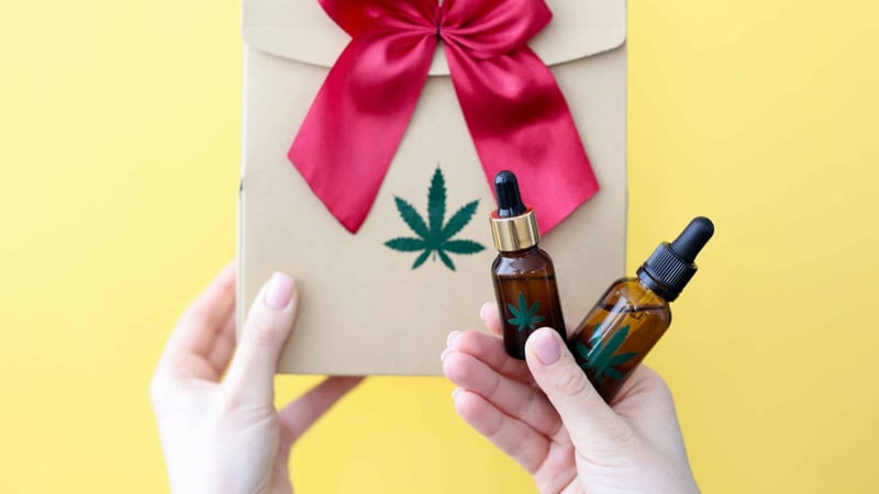 Hands Holding Two Bottles of Delta 8 Tincture and Paper Bag with Hemp Print and Pink Ribbon