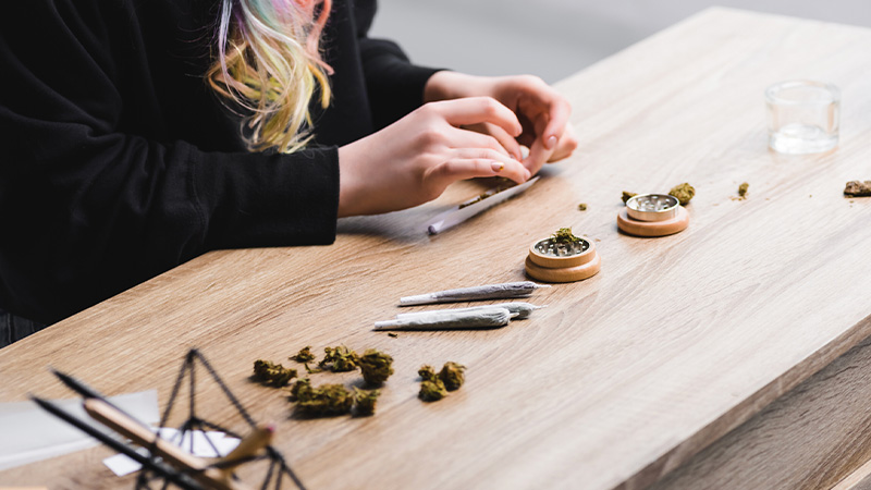 A woman rolling a joint with a grinder, rolling paper and marijuana